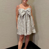 Woman wearing a floral dress with a large bow standing against a plain wall.