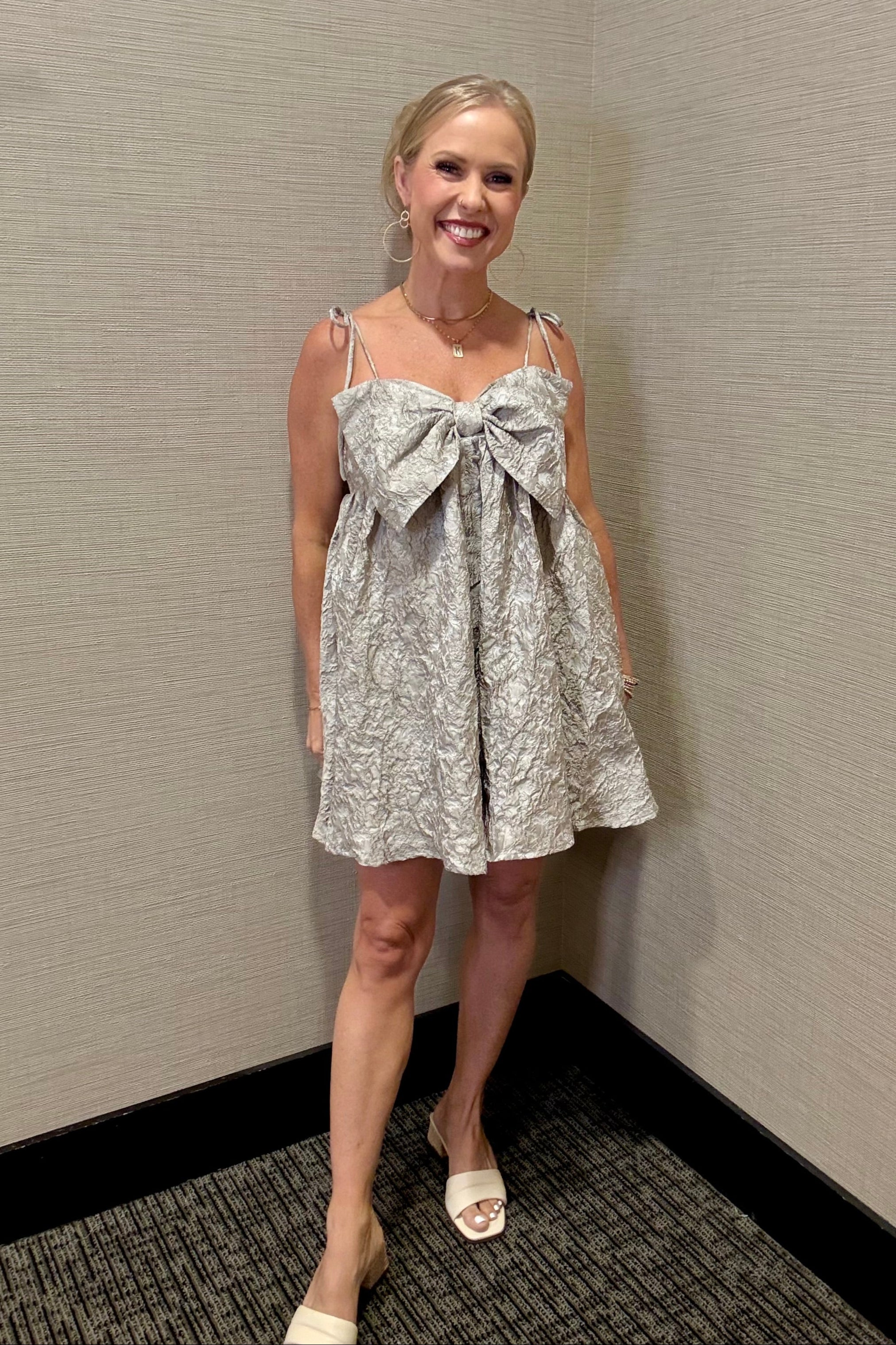 Woman wearing a floral dress with a large bow standing against a plain wall.