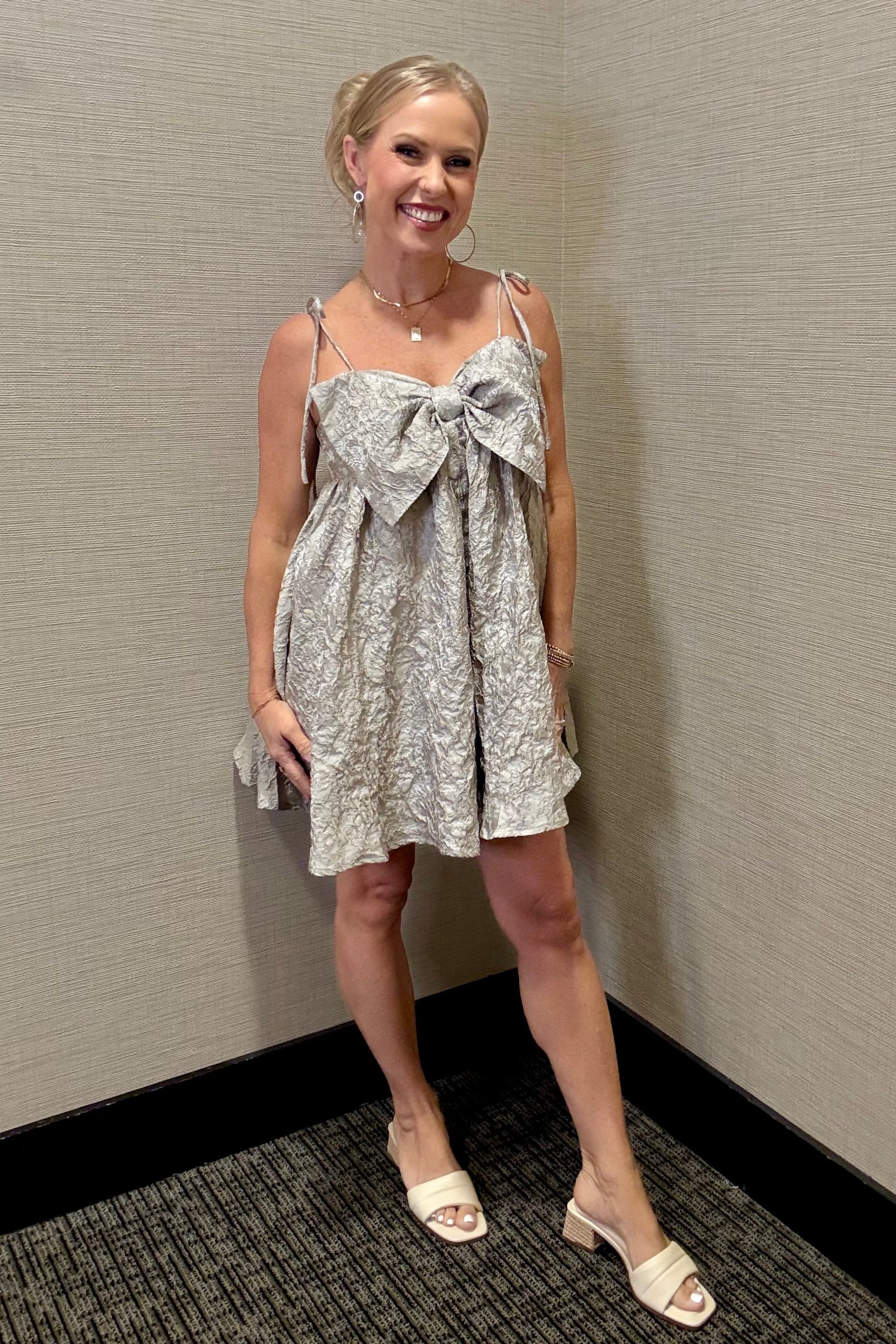 Woman wearing a patterned dress with a bow standing against a plain wall.