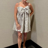 Woman wearing a patterned dress with a bow detail against a plain wall.