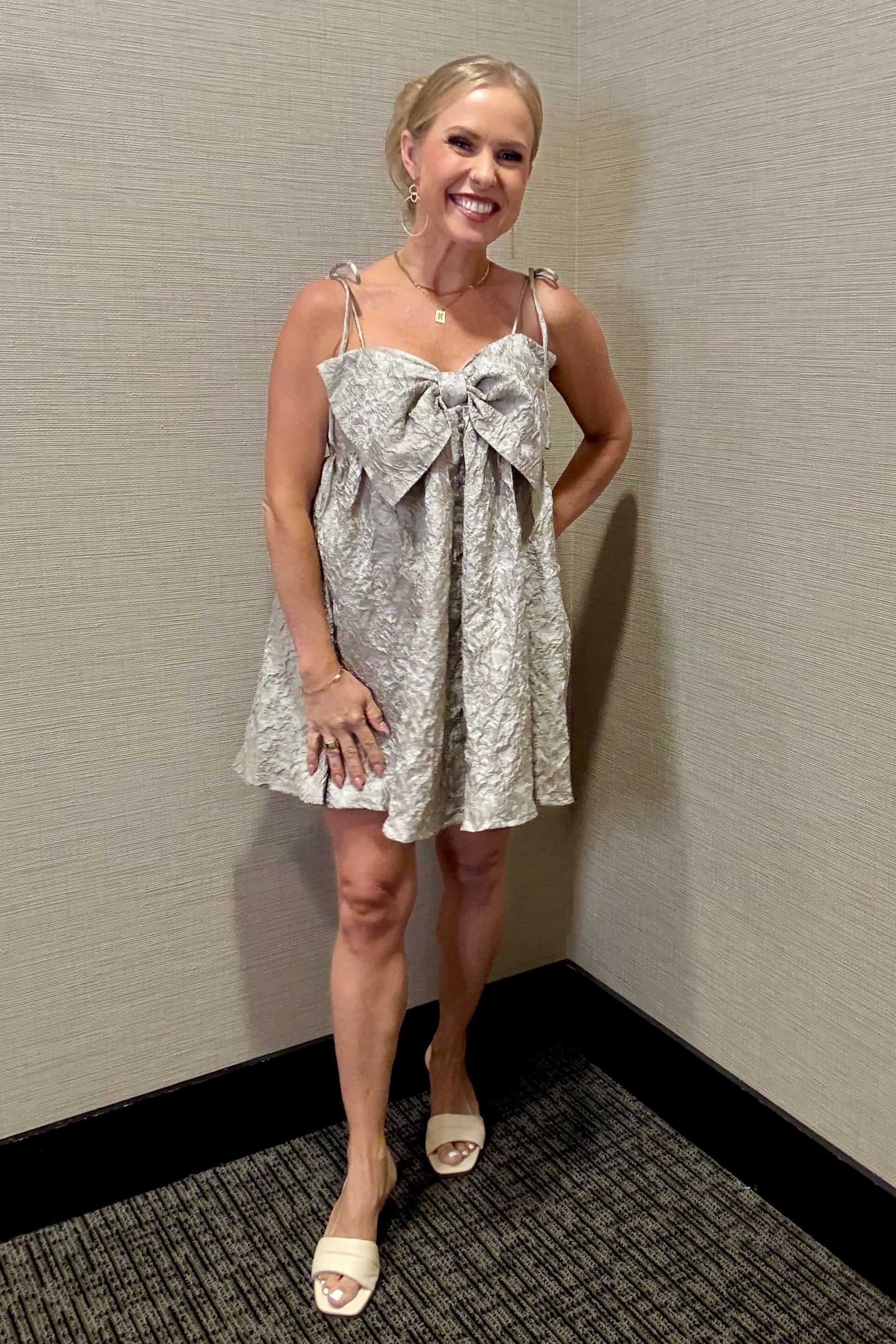 Woman wearing a patterned dress with a bow detail against a plain wall.