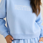 Woman wearing a light blue 'RHYTHM' sweatshirt against a beige background