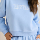 Woman wearing a light blue 'RHYTHM' sweatshirt against a beige background