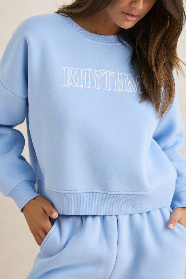 Woman wearing a light blue 'RHYTHM' sweatshirt against a beige background