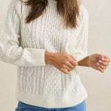 Woman wearing a white cable knit sweater and blue jeans against a beige background