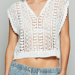 Woman wearing a white crochet top and blue pants against a gray background