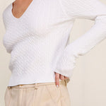 Woman wearing a white textured sweater and beige pants against a plain background