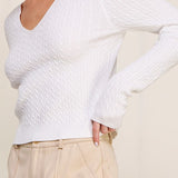 Woman wearing a white textured sweater and beige pants against a plain background