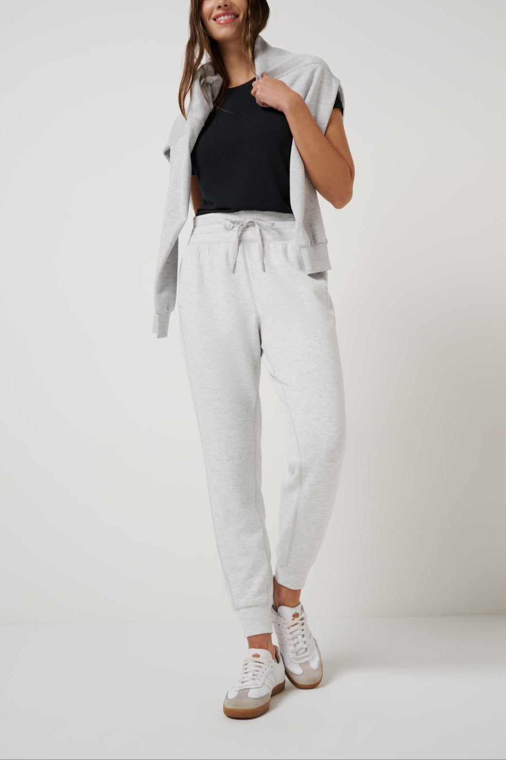 Woman wearing a black top, light gray cardigan, white pants, and white sneakers on a white background