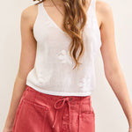 Woman wearing a white sleeveless top with floral details and red pants against a plain background