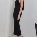 Woman in a black dress standing in a minimalistic room with a plant and chair.