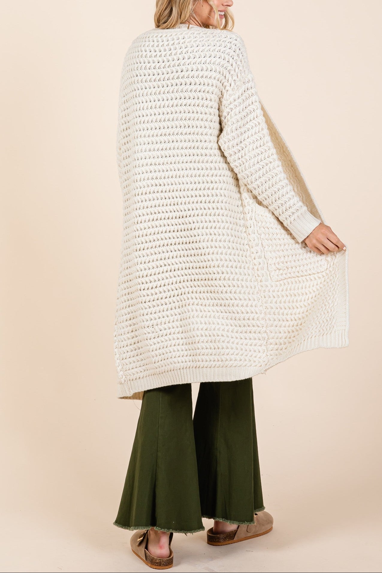 Woman wearing a long cream cardigan over green pants on a beige background