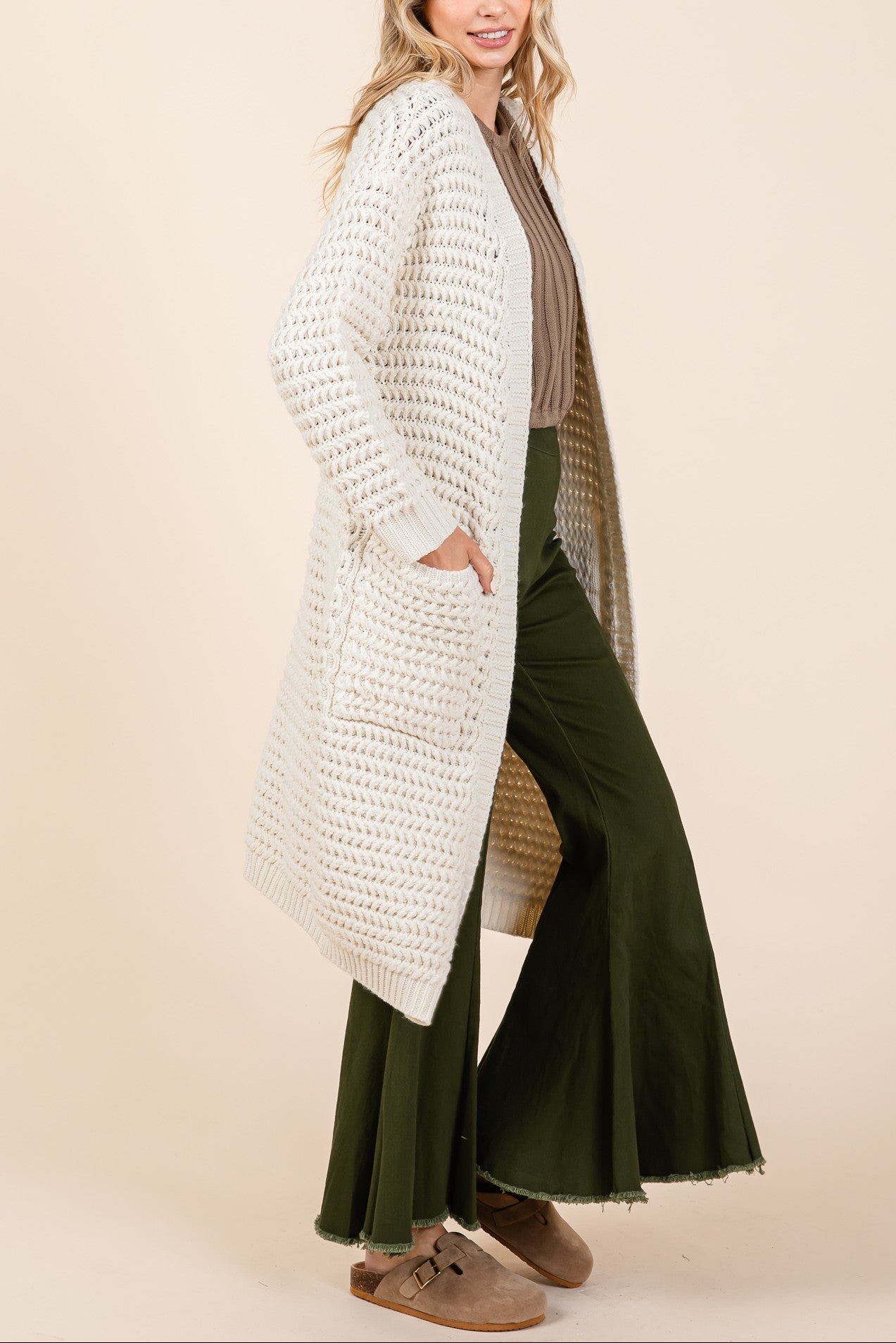 Woman wearing a long white cardigan over a beige top and green pants on a beige background