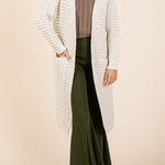 Woman wearing a long cream cardigan over a brown top and green pants on a beige background