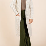 Woman wearing a long cream cardigan over a brown top and green pants on a beige background