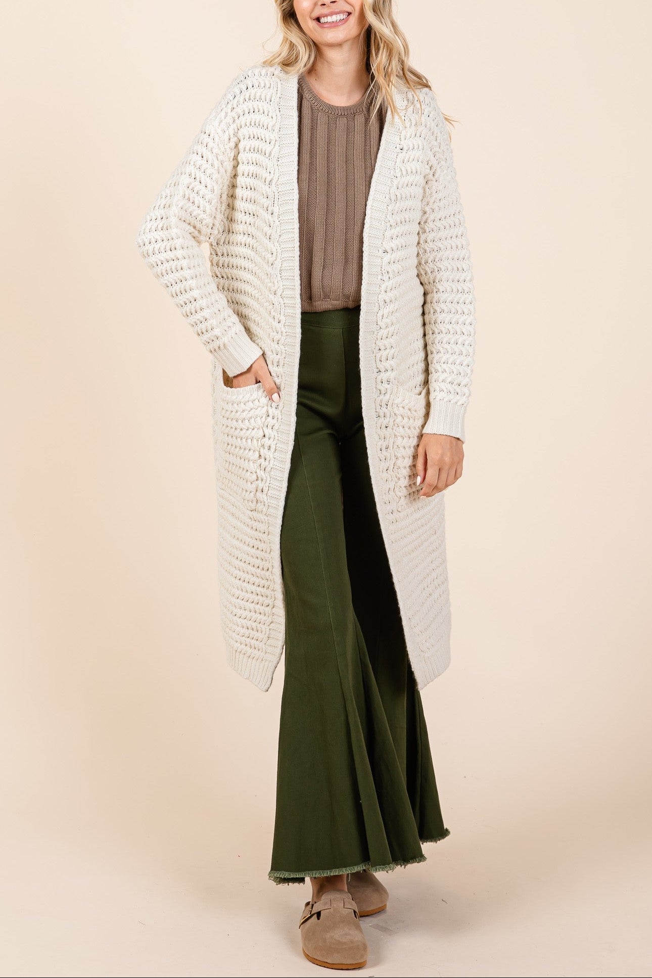 Woman wearing a long cream cardigan over a brown top and green pants on a beige background