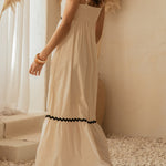 Woman in a long white dress with black trim standing in a softly lit room with decorative plants.