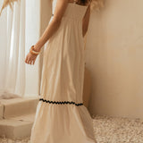 Woman in a long white dress with black trim standing in a softly lit room with decorative plants.