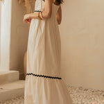 Woman in a white dress with black trim standing in a softly lit room with decorative plants.