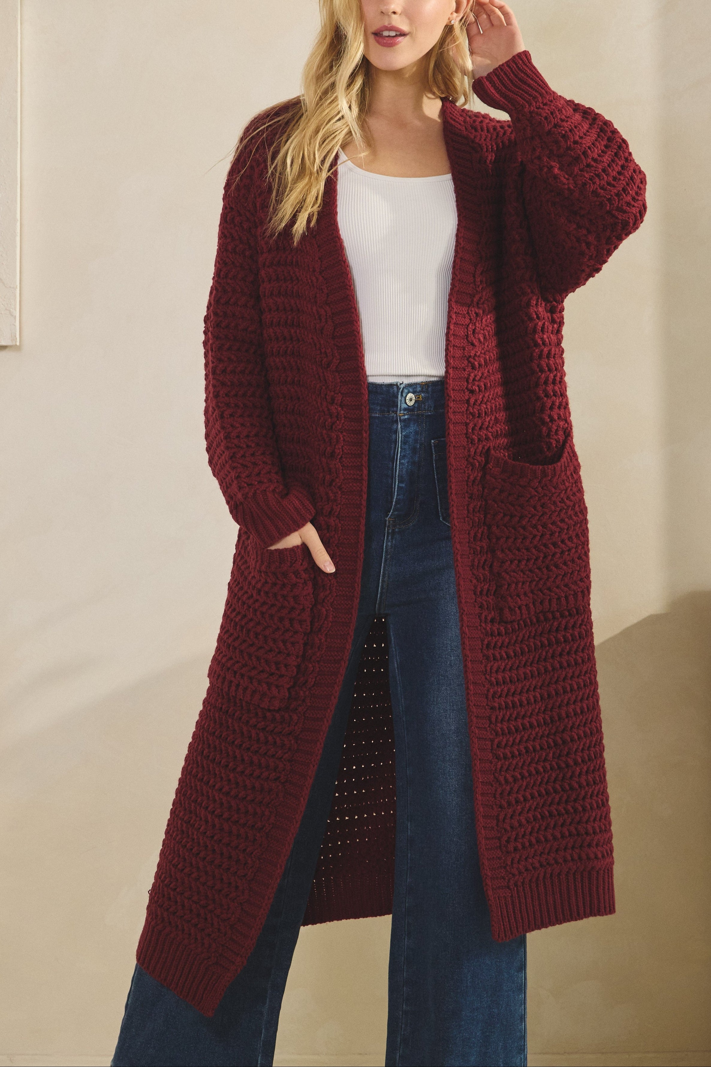 Woman wearing a long burgundy cardigan over a white top and blue jeans indoors.