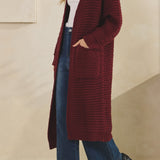 Woman wearing a long burgundy cardigan over a white top and blue jeans, standing against a beige wall.