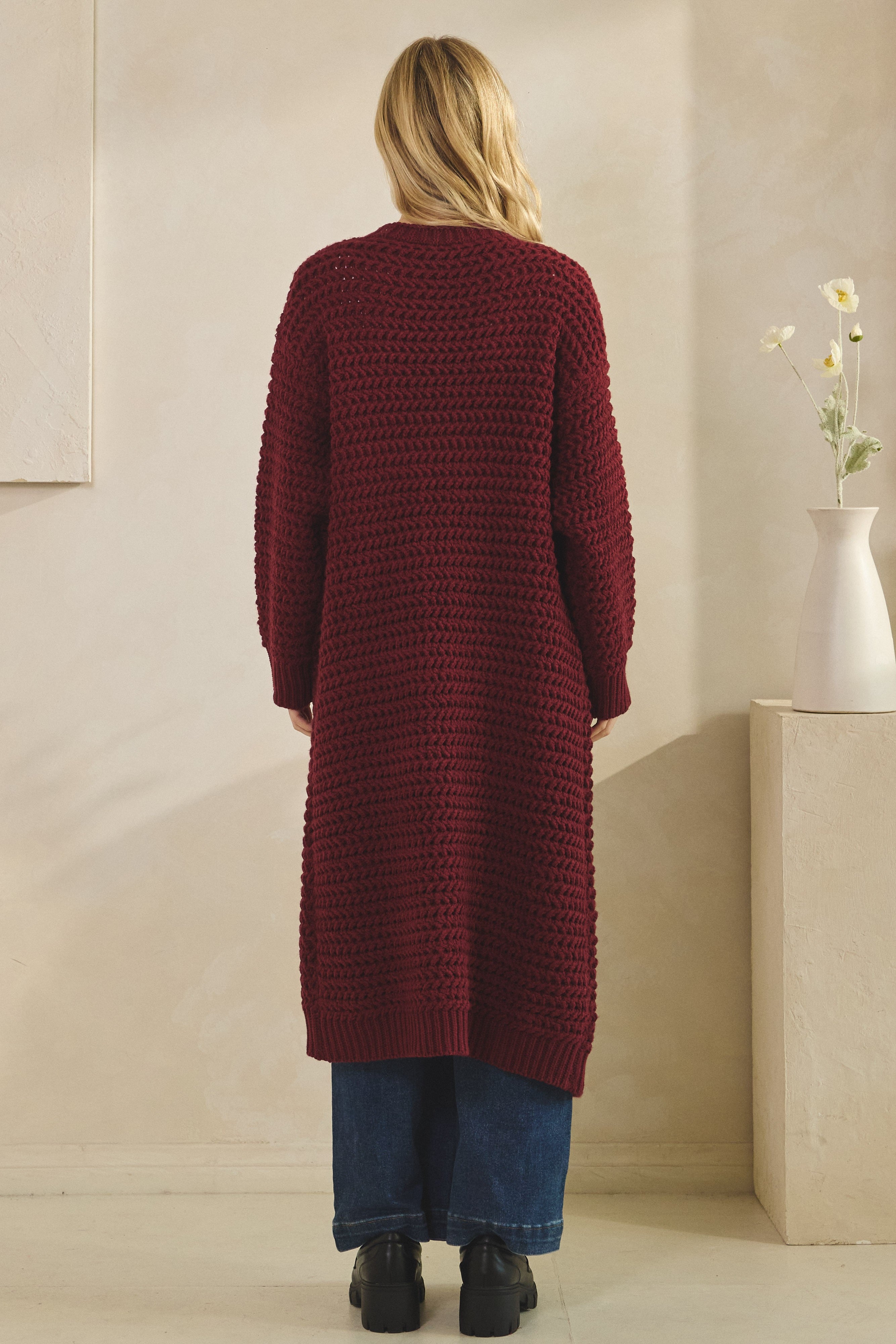 Person wearing a long burgundy cardigan in a minimalistic room.