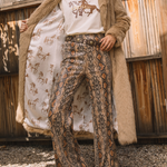 Person wearing a long beige coat, white shirt with graphic design, and patterned pants outdoors.