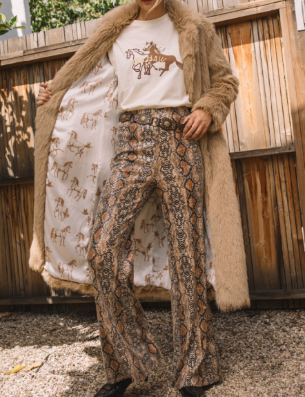 Person wearing a long beige coat, white shirt with graphic design, and patterned pants outdoors.