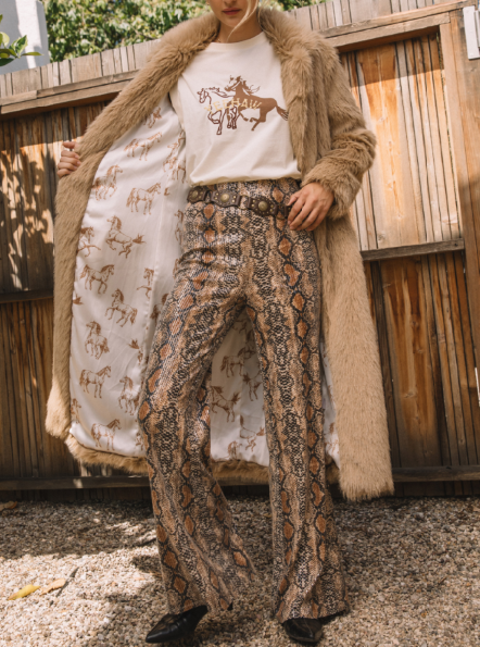 Person wearing a long beige coat, white shirt with graphic design, and patterned pants outdoors.