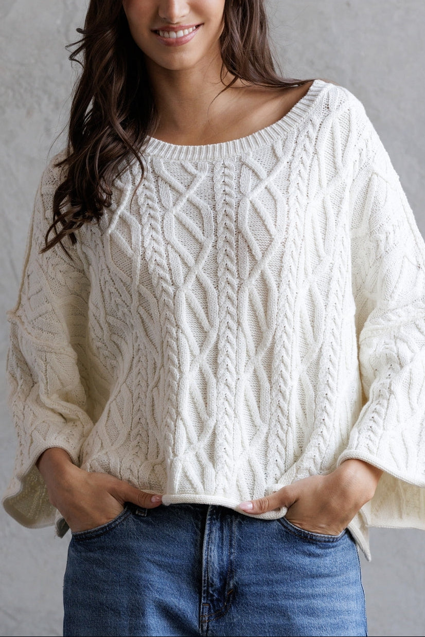 Person wearing a white cable knit sweater and blue jeans against a gray background