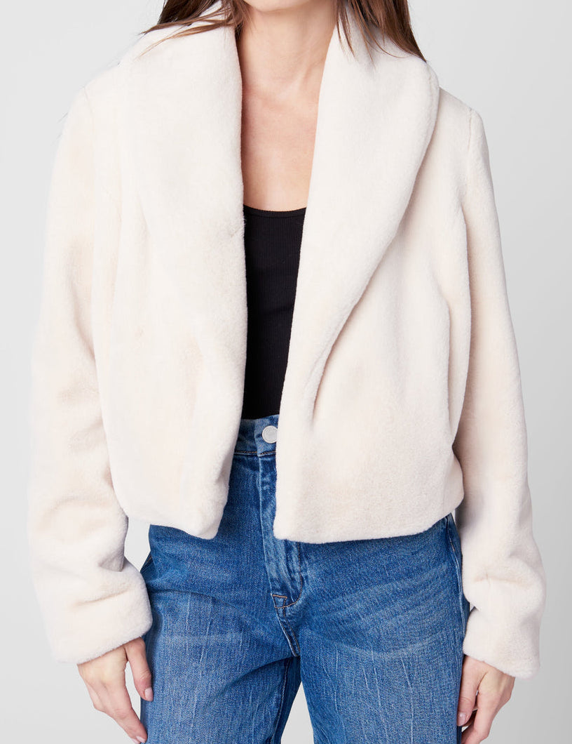 Person wearing a cream jacket over a black top and blue jeans on a white background