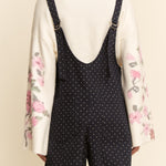 Person wearing a white shirt with floral sleeves and black polka dot overalls on a beige background