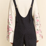 Person wearing a white shirt with floral sleeves and black polka dot overalls on a beige background