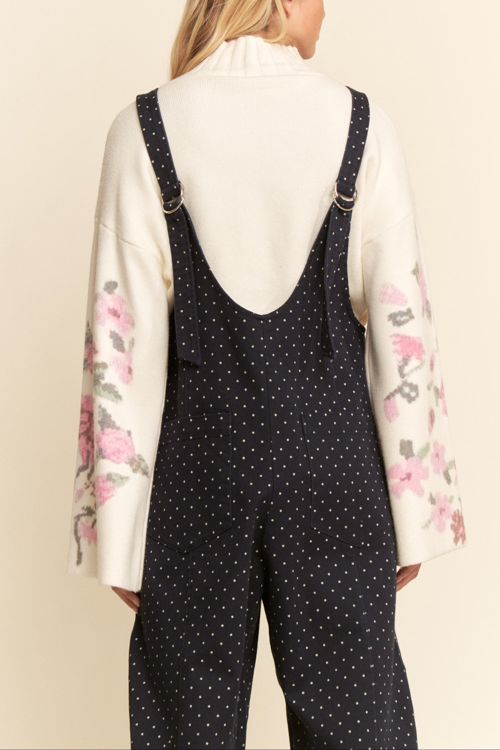 Person wearing a white shirt with floral sleeves and black polka dot overalls on a beige background