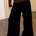 A person wearing black wide-leg pants and a brown long-sleeve top, standing indoors on a wood floor in front of a light wood slat wall.