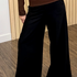 A person wearing black wide-leg pants and a brown long-sleeve top, standing indoors on a wood floor in front of a light wood slat wall.