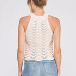 Woman wearing a white sleeveless knitted top and blue jeans on a light gray background