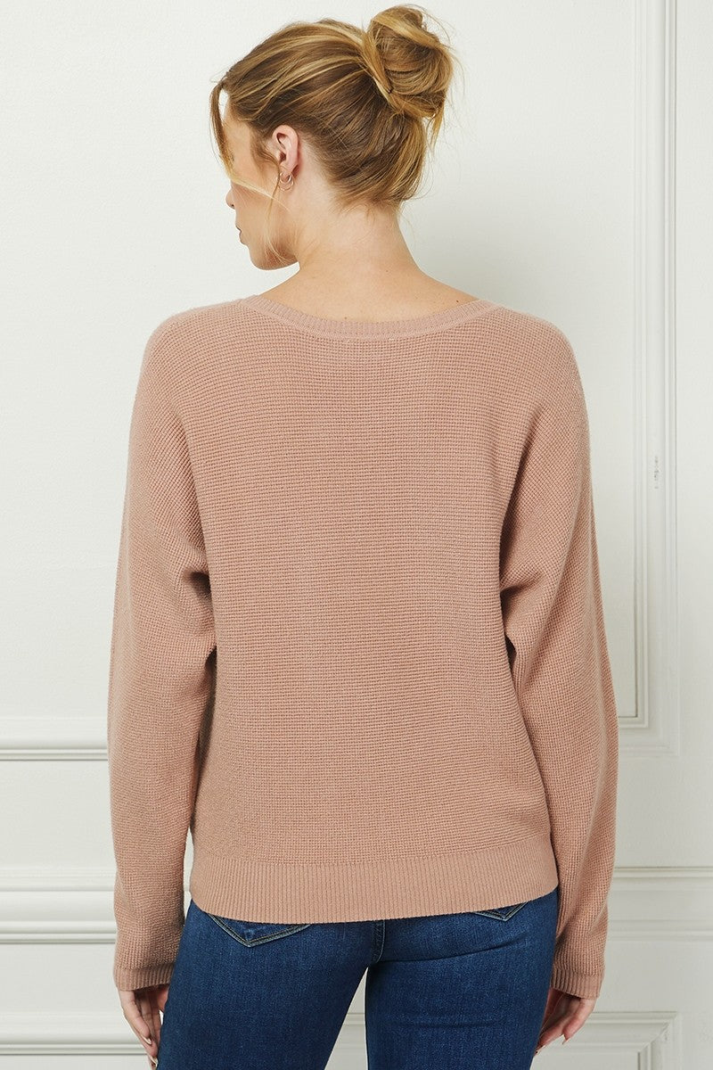 Woman wearing a beige sweater and blue jeans against a white wall.