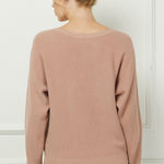 Woman wearing a beige sweater and blue jeans against a white wall.