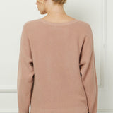 Woman wearing a beige sweater and blue jeans against a white wall.