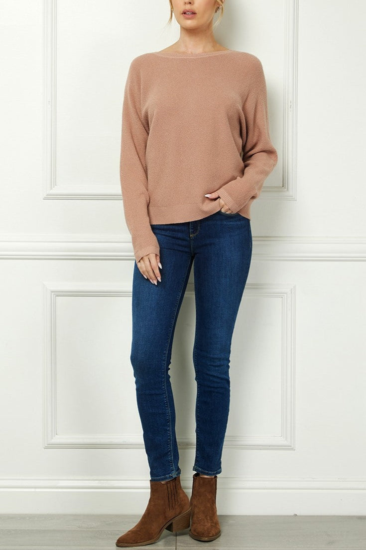 Woman wearing a beige sweater, blue jeans, and brown boots against a white paneled wall.