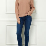 Woman wearing a beige sweater, blue jeans, and brown boots against a white paneled wall.