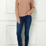 Woman wearing a beige sweater, blue jeans, and brown boots against a white paneled wall.