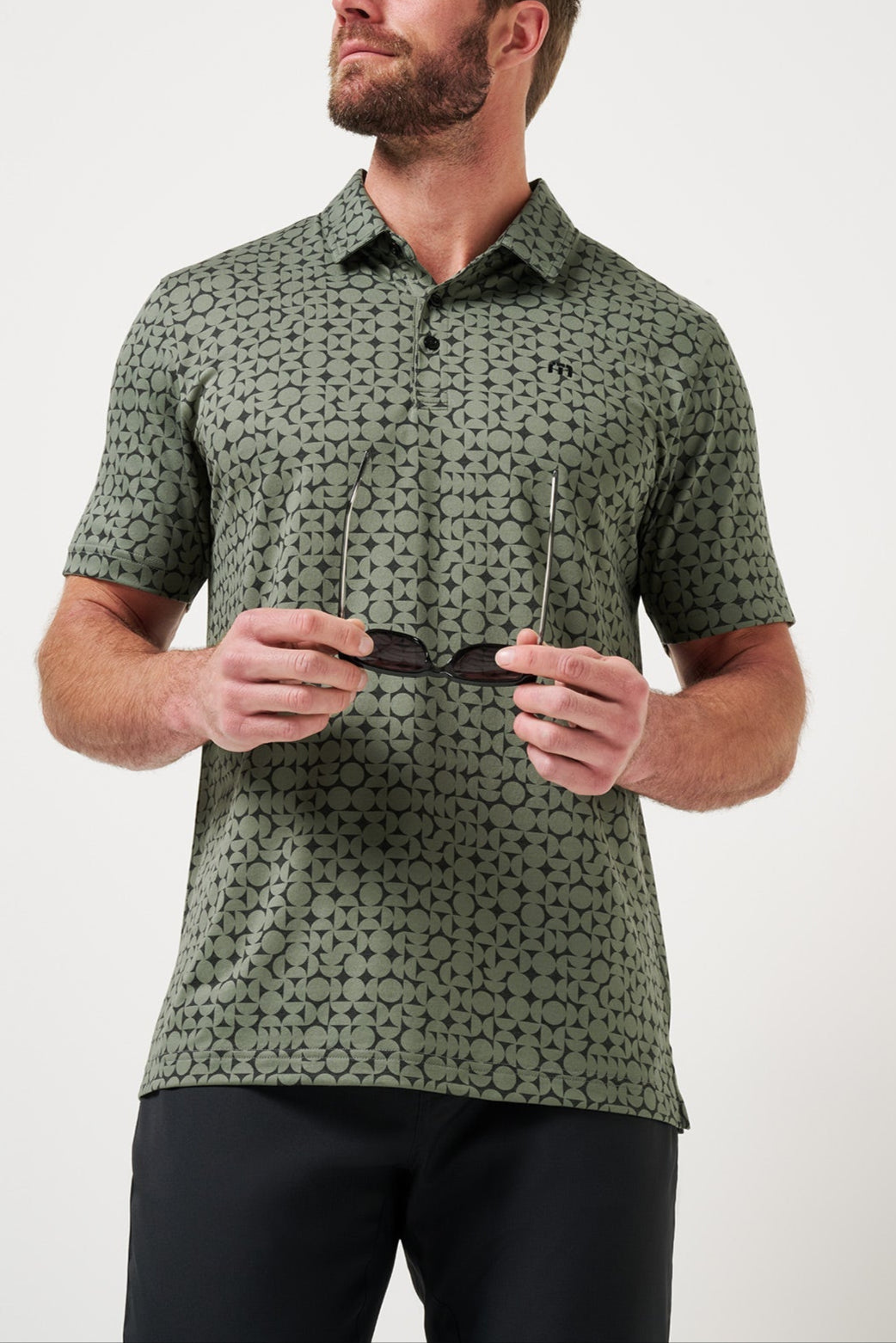 Man wearing a green patterned shirt holding sunglasses against a white background