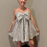 Woman wearing a floral dress with a large bow standing against a plain wall.