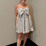 Woman wearing a floral dress with a large bow standing against a plain wall.