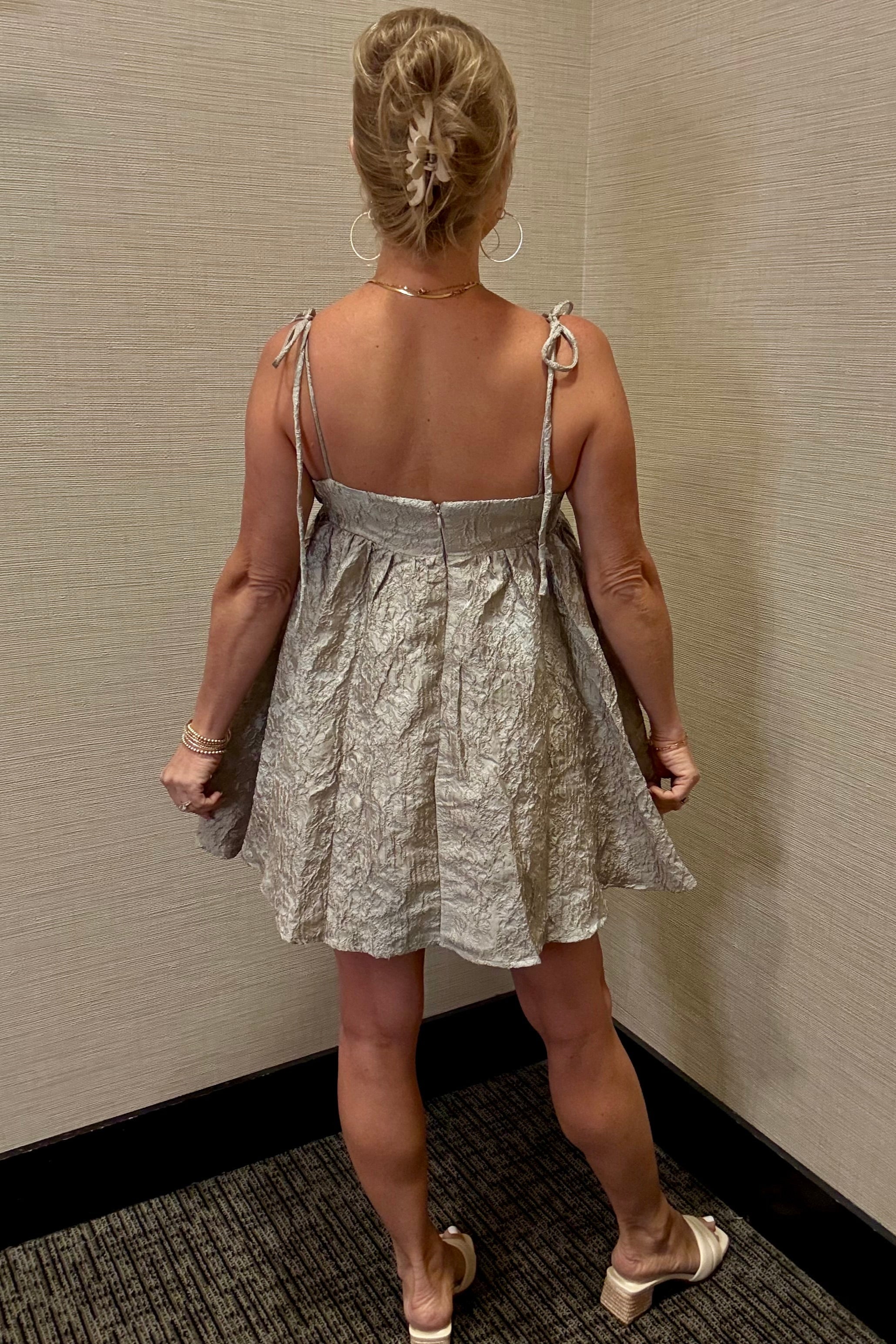 Woman wearing a silver dress with thin straps against a beige wall.