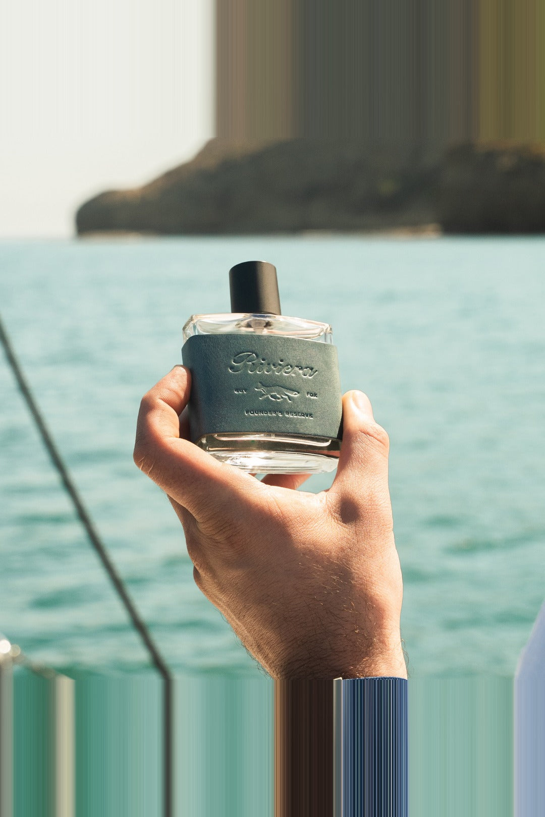 Hand holding Guy Fox Riviera cologne bottle above the water on a boat