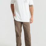 Man wearing white Roark t-shirt with small logo and brown pants on gray background