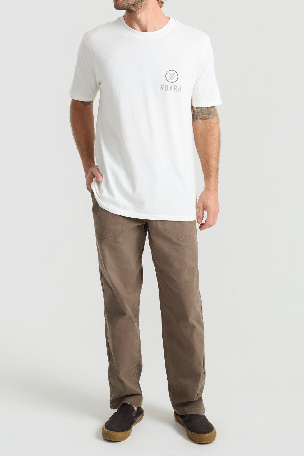Man wearing white Roark t-shirt with small logo and brown pants on gray background